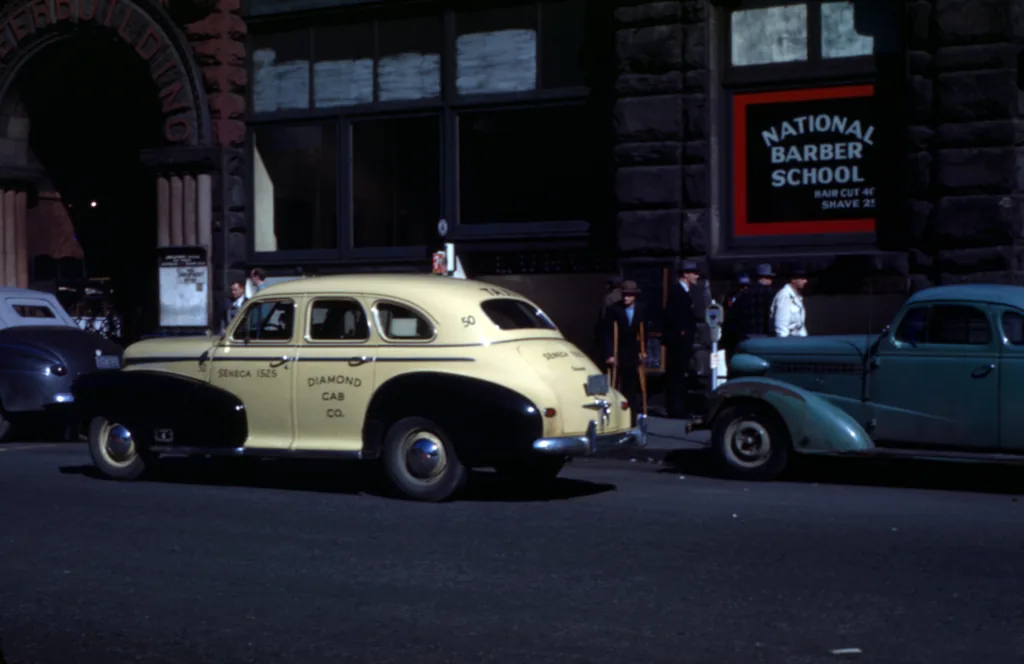 A cream Diamond Cab parked on a Pioneer Square street in Seattle, a National Barber School sign behind it and two pre-war cars alongside.
