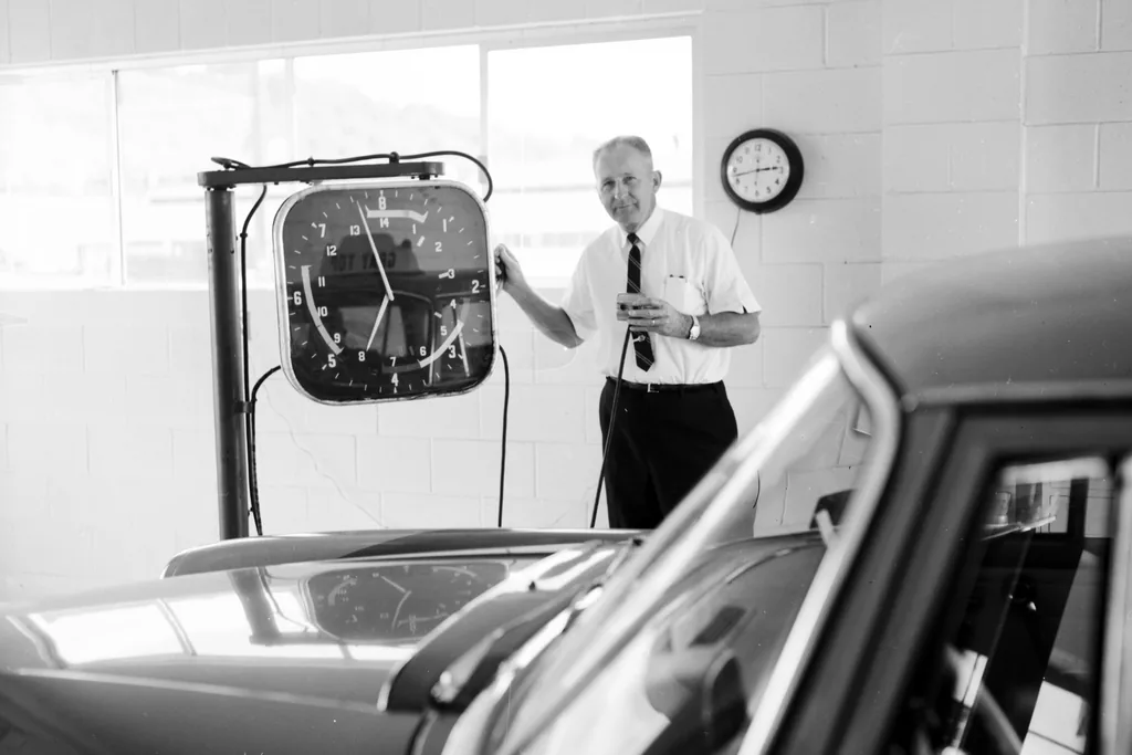 An inspector in shirt and tie stands beside a wall-mounted headlamp test rig, a car pulled up in front of him inside a municipal testing station.