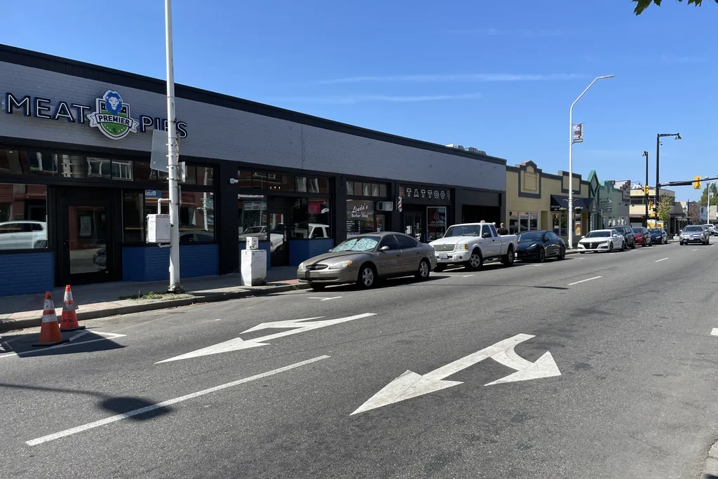 A commercial arterial in Renton on a clear day, painted turn arrows on the asphalt and parked cars along a low row of shopfronts.